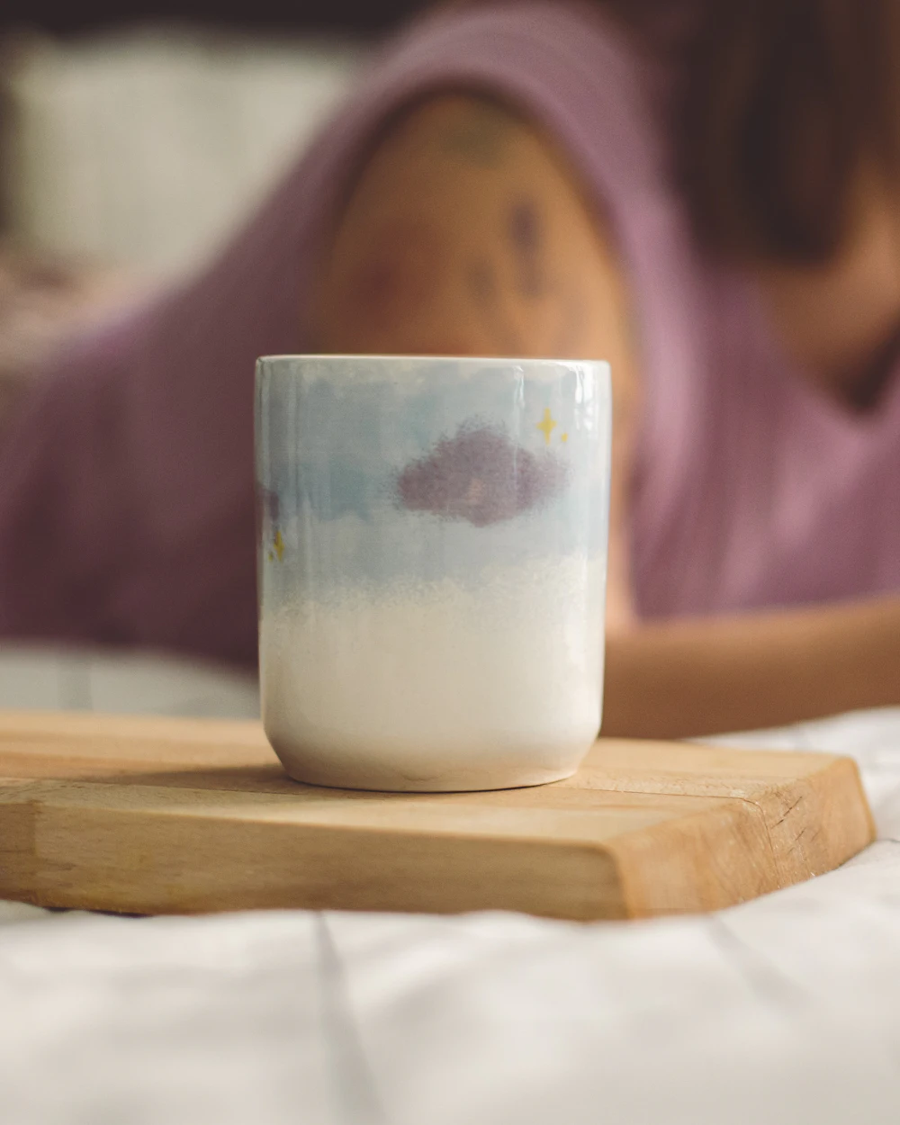 A ceramic mug on a wooden board beside an unmade bed in morning light